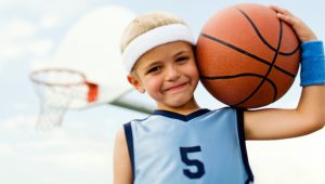 A child is playing sports. He is wearing a blue basketball tee with the number 5 on it. He has a white headband on. He is carrying a basketball next to his face/